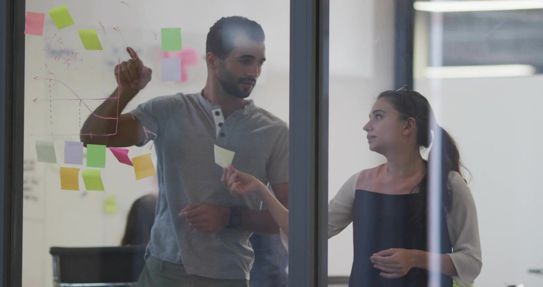 Two colleagues collaborating on glass wall with sticky notes and hand-drawn charts