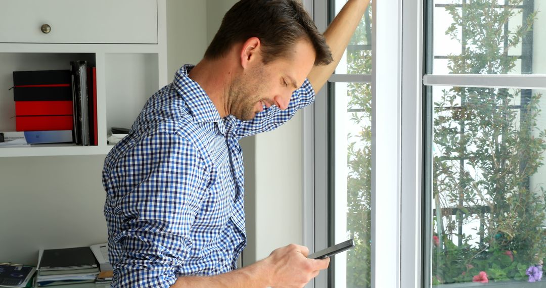 Man in casual attire standing by window looking at smartphone - Free Images, Stock Photos and Pictures on Pikwizard.com