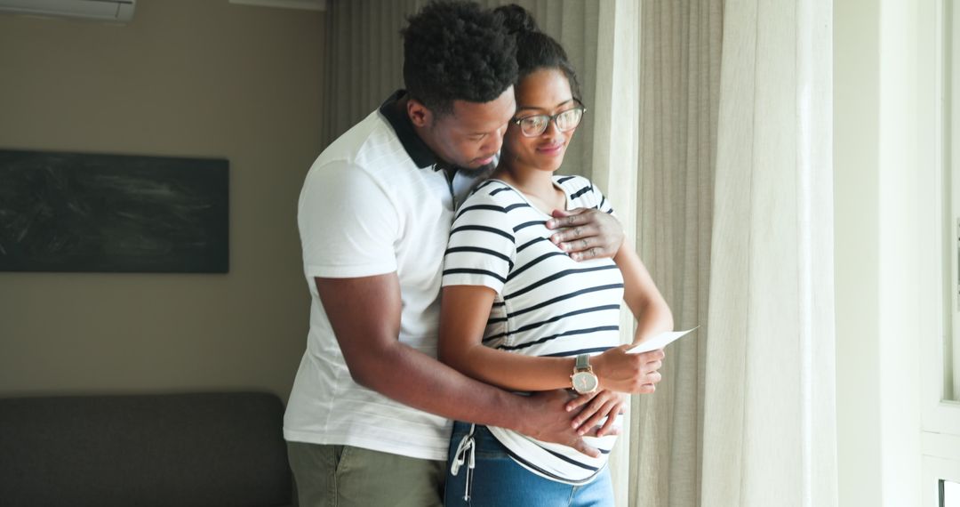 Expectant Couple Embracing Joyfully at Home by Window