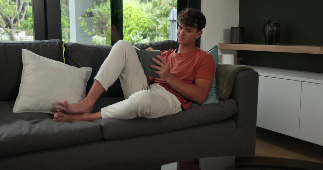Young man reclining on sofa using tablet in modern living room with garden view