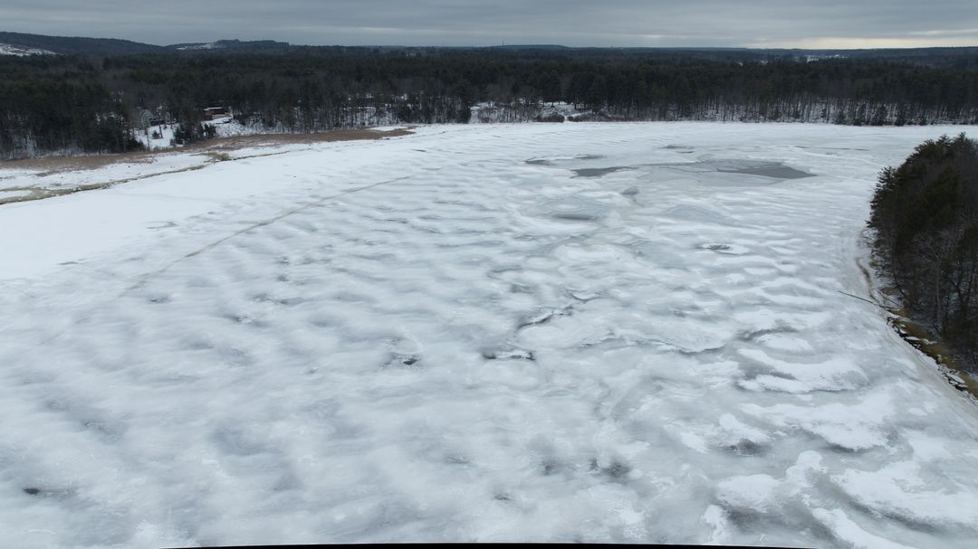 Aerial view showing frozen lake with rippled snow, winter shoreline and distant forest