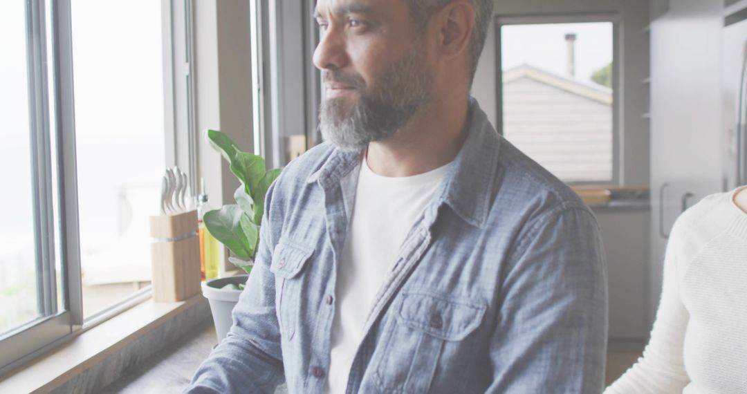 Bearded man gazing out kitchen window with partner at sunlit countertop, minimalist home