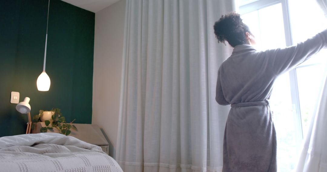 Back of biracial woman in bathrobe opening curtains in bedroom, copy space - Free Images, Stock Photos and Pictures on Pikwizard.com