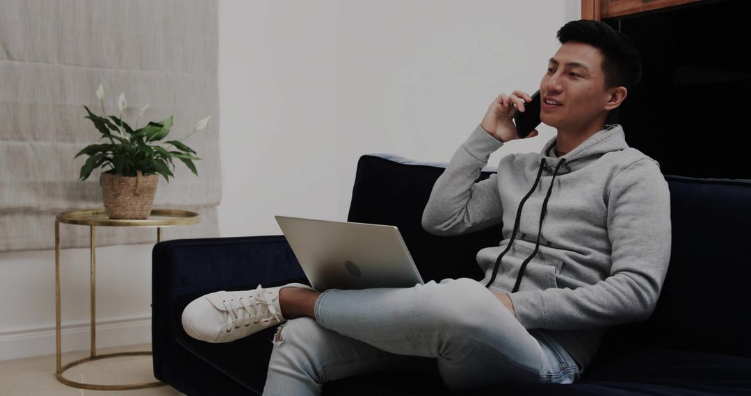 Asian man working from home on laptop while talking on smartphone on sofa