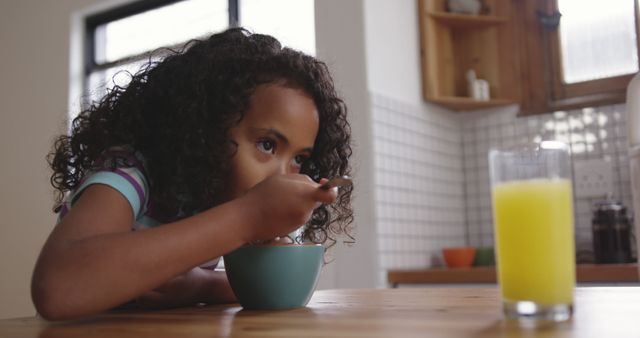 Young girl with curly hair enjoying breakfast while sitting at a wooden kitchen table. She is focused on eating from a bowl with a spoon, and a glass of orange juice is placed nearby. This can be used in contexts related to healthy eating, morning routines, family life, childhood nutrition, and home lifestyle settings.