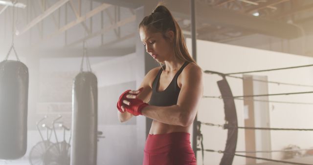 Female boxer wrapping her hands with red wraps in a gym before a workout. Ideal for use in fitness blogs, boxing training programs, women's sports promotional materials, and motivational posters.