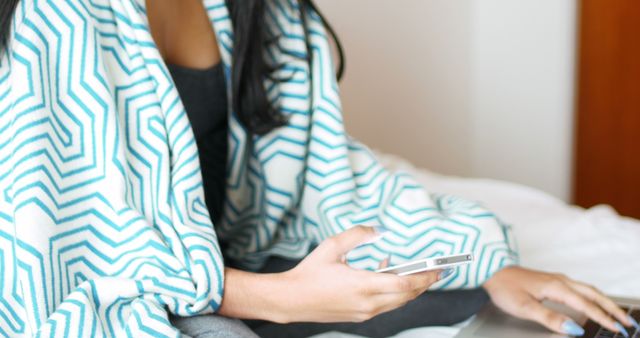 Woman covered with stylish blanket using smartphone and laptop while sitting at home. Ideal for technology, lifestyle, remote work, and relaxation themes.