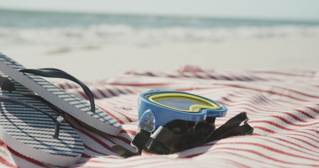 Capture the essence of summer relaxation with this image of striped flip-flops and a pair of goggles resting on a red and white striped blanket by the ocean. Ideal for travel blogs, vacation promotion, summer merchandise, and coastal lifestyle content.