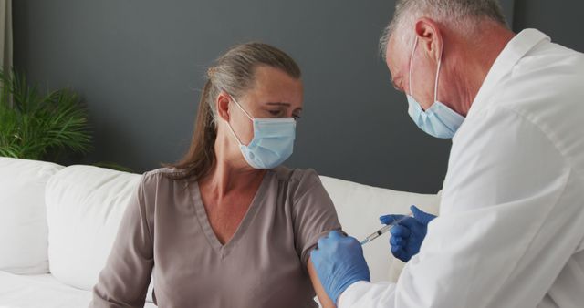 Mature woman receiving vaccine from healthcare professional at home. Both are wearing masks for protection. This can be used for health-related campaigns, articles about vaccination drives, elderly care, home healthcare services, and COVID-19 safety measures.
