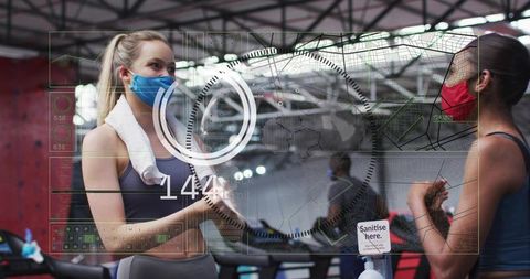 Women wearing masks talking at gym near sanitizer with futuristic hud fitness technology