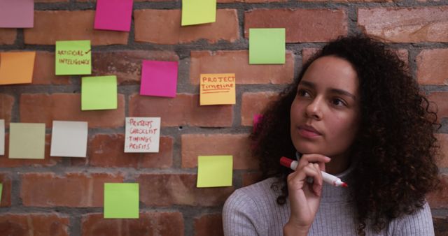 Young biracial professional looking at colorful sticky notes on brick wall. She has curly brown hair, light brown skin, and is wearing a casual top