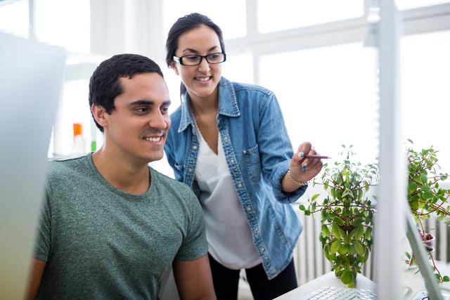 This image shows two graphic designers working together in a modern office environment. The male designer is sitting at a computer while the female designer is standing and pointing at the screen, indicating a collaborative discussion. Both are dressed in casual attire, and the office is decorated with plants, adding a touch of greenery. This image can be used for articles or websites related to teamwork, creative industries, office culture, or design projects.
