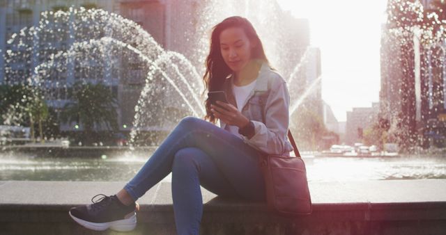 Asian woman sitting next to fountain using smartphone and drinking takeaway coffee. digital nomad on the go, out and about in the city.