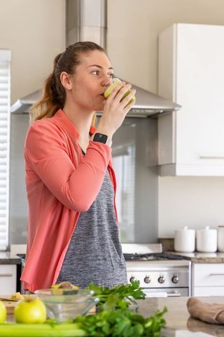 Young woman sipping green smoothie in modern kitchen wearing coral cardigan and smartwatch