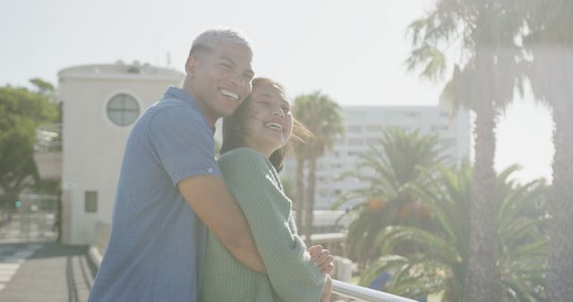 A joyful couple is embracing outdoors on a sunny day with a scenic backdrop of palm trees and a modern building. Suitable for use in advertisements, travel blogs, promotional materials for holiday destinations, love and relationship articles, and health and wellness campaigns celebrating moments of happiness and relaxation.