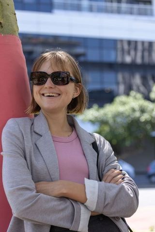 Young woman leaning on red-wrapped tree smiling wearing sunglasses and blazer urban confidence