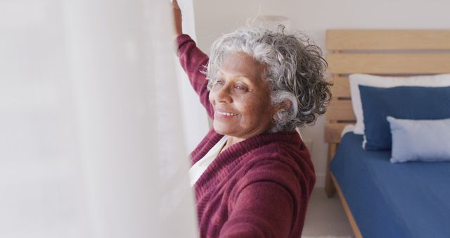 Senior woman joyfully opening curtains to let in natural light in a cozy bedroom. Suitable for promoting products or services related to senior living, healthy lifestyle for elderly, and morning routines. Can be used in articles, advertisements, and social media posts about daily habits, home decor, and wellbeing.