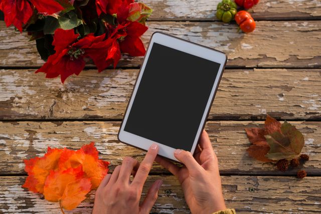 Hands interacting with a tablet on a rustic wooden table surrounded by autumn leaves and poinsettia flowers. Ideal for themes related to technology, autumn, digital interaction, and seasonal settings. Suitable for blogs, websites, and advertisements focusing on fall activities, tech usage, or rustic aesthetics.