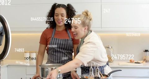 Two women cooking together in modern kitchen with aprons and ring light for streaming