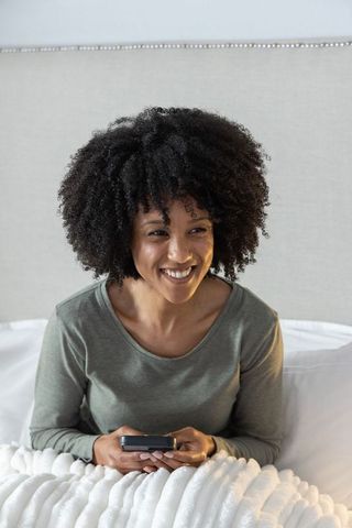 African american woman smiling, holding smartphone, sitting on bed in cozy bedroom
