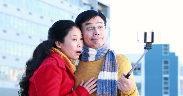 A middle-aged Asian couple looks surprised while taking a selfie with a smartphone on a selfie stick, with copy space. Their playful expressions add a sense of fun and spontaneity to the moment captured.