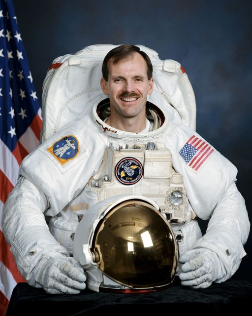 American astronaut in full spacesuit is holding helmet while posing in front of American flag and blue background. Ideal for educational content, articles on space exploration, inspirational posters, technology advertisements, scientific journals, space mission documentaries, and aerospace industry marketing materials.