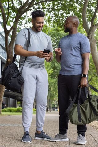 Black and latino male friends checking smartphone and holding gym duffel bags in urban park