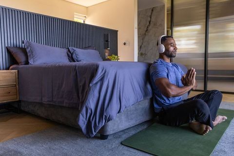 Mature african-american man meditating on yoga mat in bedroom wearing headphones