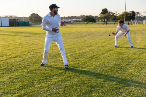 Two cricketers practicing catch and fielding on sunlit grass outfield