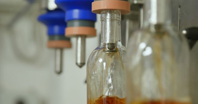 Image shows close-up of a glass bottle being filled with liquid using a bottling machine in a bottling plant. This photo can be used to illustrate automated manufacturing processes in beverage or liquid production industries, industry technology, and factory settings.