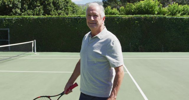 Portrait of smiling senior caucasian man holding tennis racquet on tennis court on sunny day. retirement, golden years and senior active lifestyle.