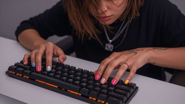 Closeup hands typing on compact mechanical keyboard with multicolor nail art and tattoos
