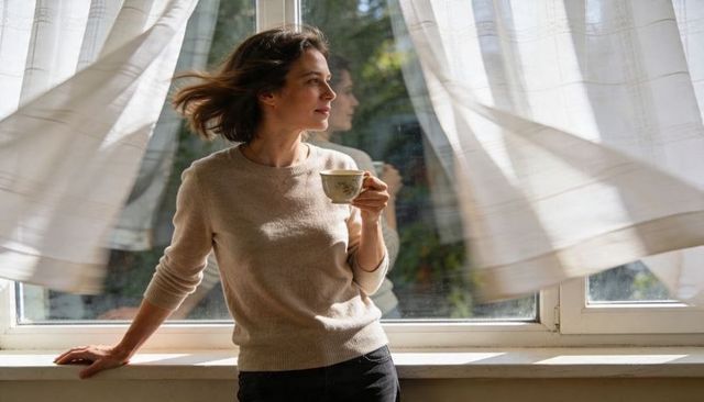 Serene morning window scene: woman gazing, holding teacup on sunlit sill with curtains