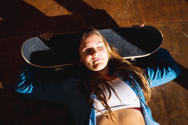 Young woman lying down with eyes closed, holding skateboard behind her head in a sunlit shop. Ideal for use in lifestyle blogs, sport and leisure advertisements, youth culture promotions, and relaxation-themed content.