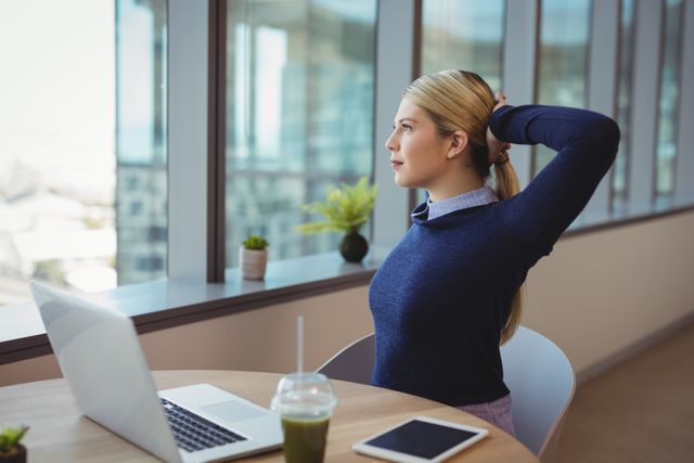 Businesswoman taking a break from work, stretching at her desk in a modern office with large windows. Ideal for illustrating workplace wellness, productivity, and healthy work habits. Can be used in articles about office ergonomics, corporate wellness programs, and work-life balance.