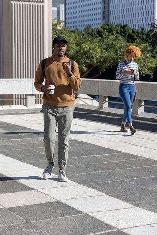African american man walking with coffee and backpack using smartphone in urban plaza