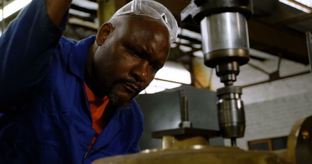 Industrial worker in blue overalls and safety glasses operating a precision machine in a factory setting. Focused expression showcasing craftsmanship and technical skill. Ideal for use in materials related to manufacturing, engineering careers, industrial training, and safety procedures.