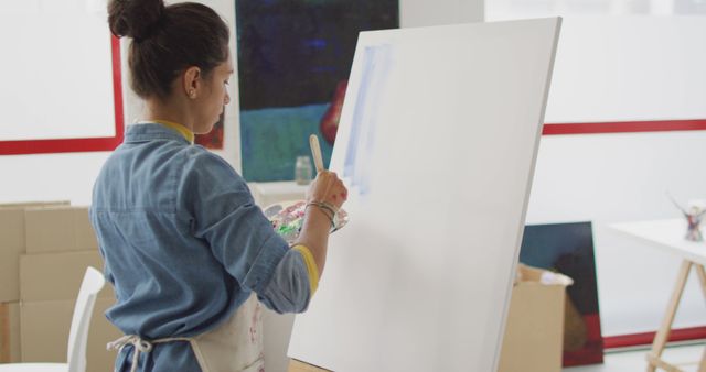 Woman artist working on large canvas in modern studio, creating new artwork using brush and palette. Ideal for use in projects about creativity, artistic process, female artists, inspiring workspaces, hobbies, and professional art.