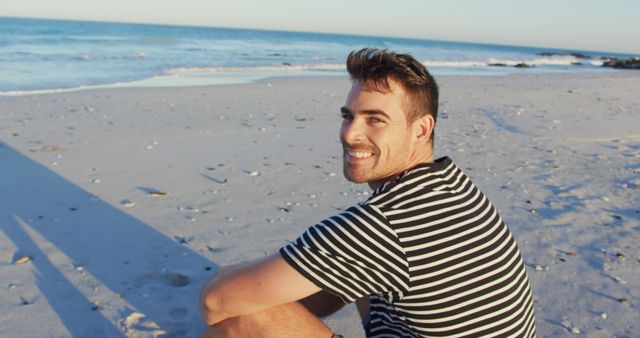 Man sitting on sandy beach during sunset, smiling and wearing a striped shirt, with the ocean in background. Ideal for use in travel promotions, leisure and vacation advertising, lifestyle blogs, and relaxation content.