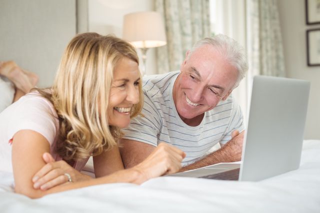 Senior couple lying on bed and using laptop, smiling and enjoying time together. Perfect for themes related to senior lifestyle, technology use among elderly, family bonding, home comfort, and leisure activities.