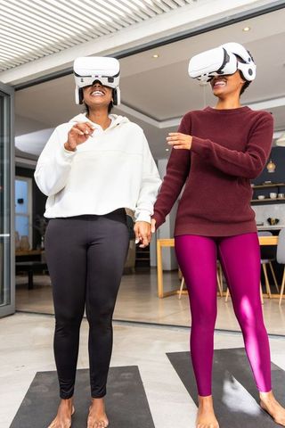 African american and indian women holding hands wearing vr headsets on yoga mats at home