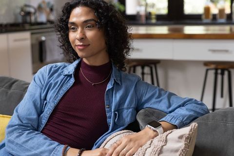 Nonbinary adult relaxing on gray sofa in open kitchen wearing denim shirt and smartwatch