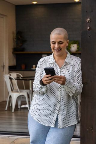 Smiling woman with shaved head leaning in doorway, checking smartphone in modern home kitchen