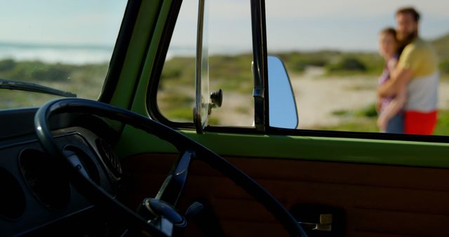 Classic car interior with a view of a couple standing on a beach outside. Ideal for themes about travel, road trips, vintage vehicles, and outdoor leisure activities. Suitable for use in travel blogs, marketing materials for vacation destinations, and articles about classic cars.