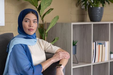 Woman wearing blue hijab sitting in modern lounge showing smartwatch for wellness and style