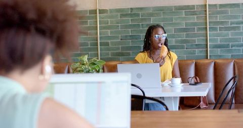 Woman in yellow top eating croissant while typing on laptop in modern cafe with green tiles