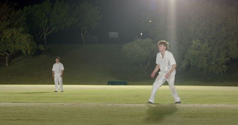 Night cricket match with bowler preparing and fielder observing on floodlit grass pitch