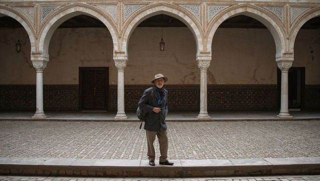 Senior traveler standing in historic arched courtyard wearing wide-brim hat and backpack