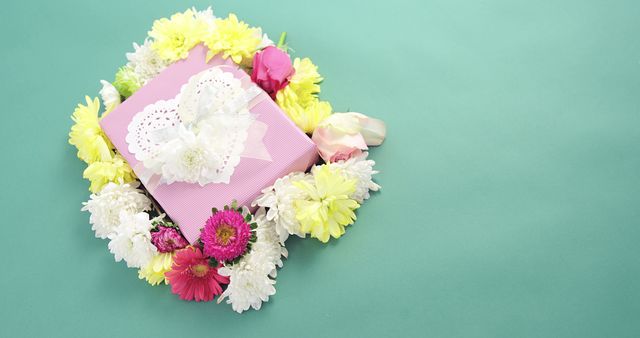 A pink gift box adorned with a lace heart and surrounded by colorful flowers rests on a mint green background, with copy space. This arrangement suggests a festive or celebratory occasion, such as Mother's Day, a birthday, or an expression of appreciation.