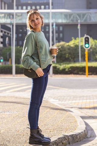 Young woman standing at crosswalk holding coffee wearing casual jeans sweatshirt and boots